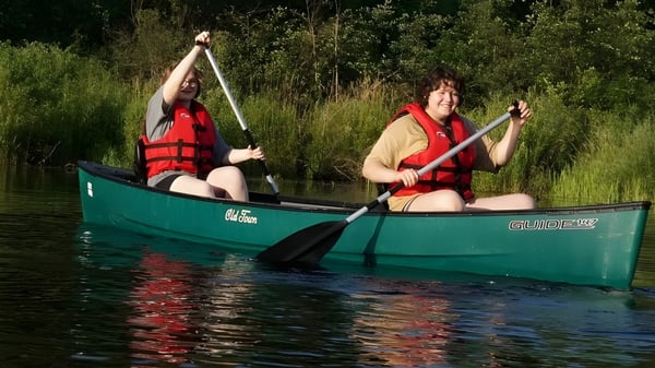 Dos alumnos de The Marvelwood School reman con chalecos salvavidas en un kayak verde en un cuerpo de agua rodeado de vegetación.