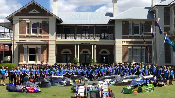 Estudiantes de la Maryborough State High School se reúnen frente a un edificio histórico junto a varios coches de carreras.