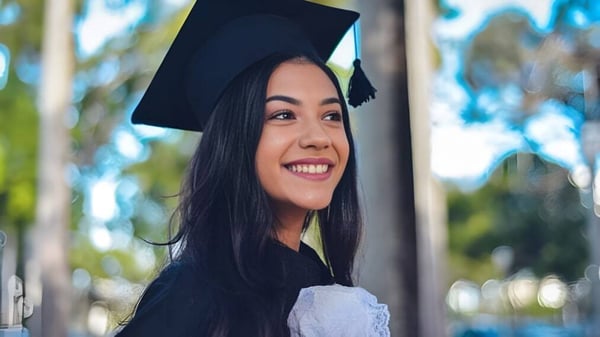 Una joven en ropa de graduación está de pie frente a un fondo natural en el campus de la Matthew Halton High School.