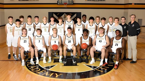 El equipo de baloncesto de la Maur Hill-Mount Academy posan junto con los entrenadores en la cancha frente al logo del equipo.