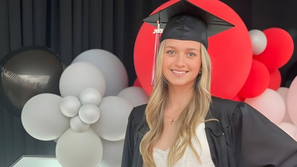 Una estudiante de la Maxwell Heights Secondary School está en su toga de graduación frente a globos rojos y blancos.