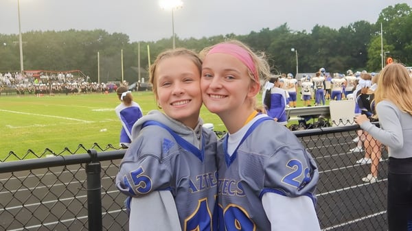 Dos jóvenes deportistas están sonriendo en el campo de deportes de la Maynard High School con espectadores al fondo.