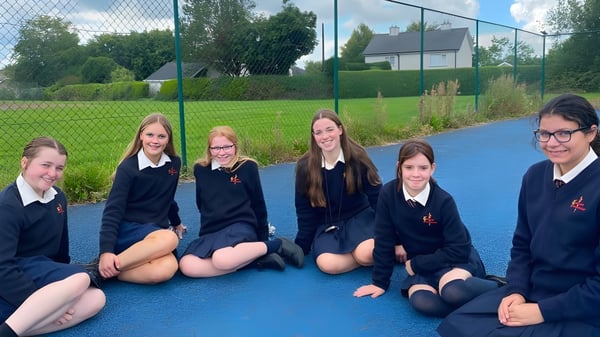 Alumnas de la Meán-Scoil Mhuire Gan Smál están sentadas en uniforme sobre una superficie de goma azul en el espacio exterior de la escuela.