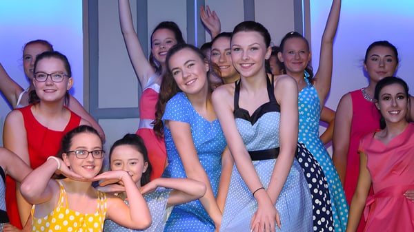 Un grupo de mujeres vestidas de manera colorida posan frente a un fondo estampado en el campus de la Meán-Scoil Mhuire Gan Smál.
