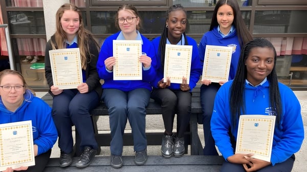 Estudiantes de la Meán Scoil Mhuire en Longford están en uniformes azules con diplomas frente a un edificio de ladrillo.