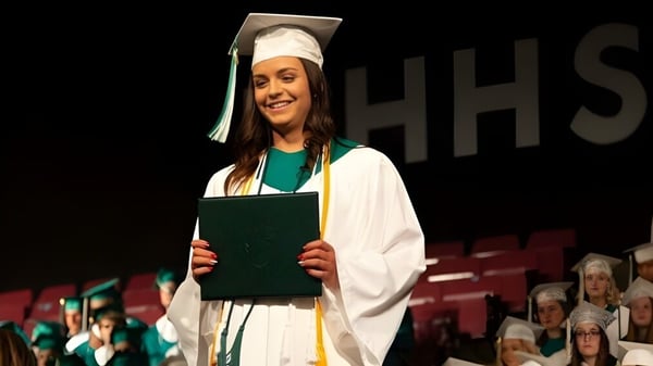 Una graduada sonriente de la Medicine Hat High School sostiene su diploma en el escenario frente al fondo con las letras HHS.