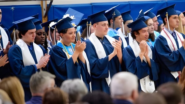 Un grupo de graduados de la Medway High School está de pie en togas azules frente a un fondo azul y mira a la cámara.