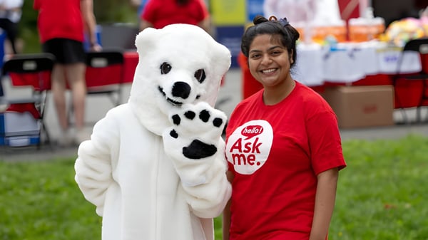 Una persona sonriente con una camiseta roja de Ask me está junto a un gran peluche blanco con huellas negras en el terreno de la Memorial University.