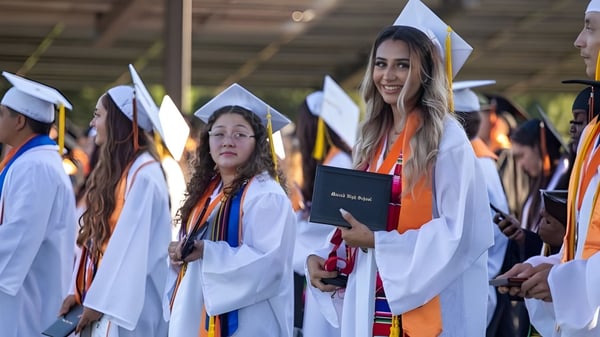 Un grupo de graduados del Merced Union High School District está en túnicas y birretes blancos con diplomas en una ceremonia.