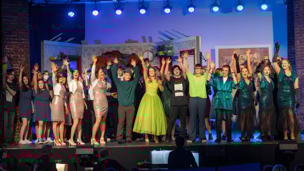 Un grupo de estudiantes de la Mercy Secondary School está en un escenario con iluminación colorida frente a un fondo de horizonte urbano.