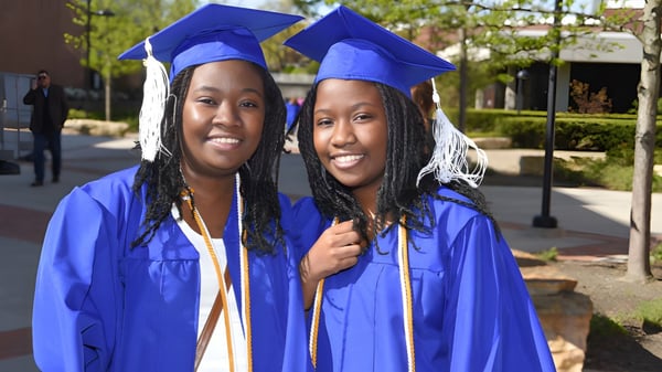 Dos estudiantes en togas de graduación azules están juntas frente a un edificio del campus del Meridan State College.