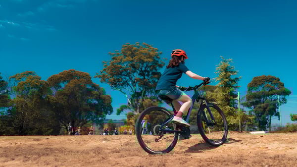 Una estudiante del Meridan State College monta una bicicleta de montaña en un sendero forestal bajo un cielo azul.