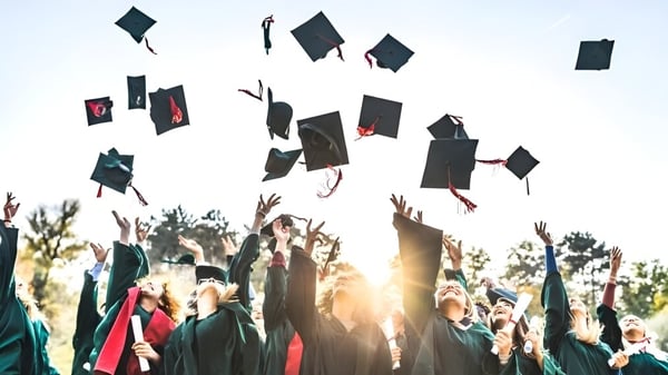 Los graduados de la Merivale High School lanzan sus birretes al aire frente a árboles y un sol poniente.