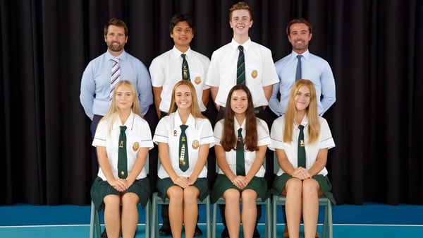 Un grupo de estudiantes de la Merrimac State High School está sentado en uniformes escolares frente a un fondo oscuro en una pose formal.