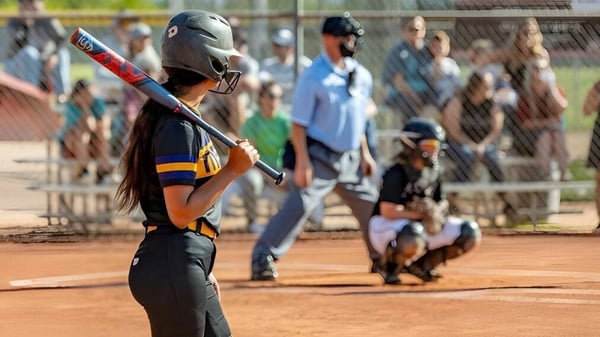 Una jugadora de softball está en el campo del Mesa Unified School District con otros jugadores y espectadores al fondo.