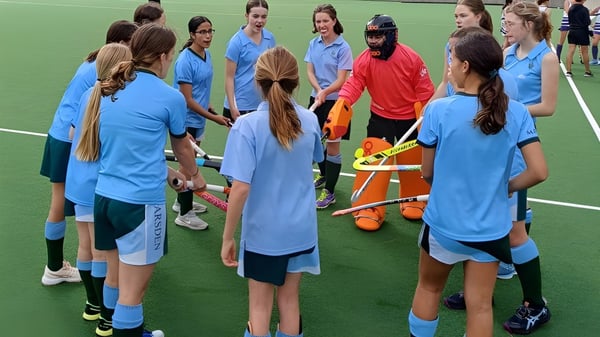 Un grupo de jóvenes deportistas en camisetas azules está en el campo de deportes del Methodist Ladies' College alrededor de una jugadora en camiseta roja.