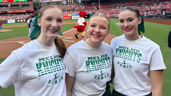 Tres estudiantes de la Metro East Lutheran High School posan sonriendo en un estadio de béisbol con camisetas blancas.