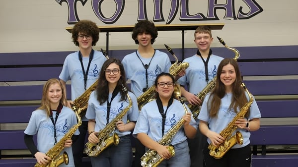 Un grupo de estudiantes de la Michigan Lutheran High School está en uniformes uniformes con saxofones frente al gran cartel de Tomcats.