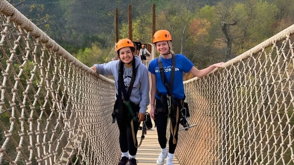 Dos personas con cascos cruzan un puente colgante en el área boscosa del terreno de la Michigan Lutheran High School.