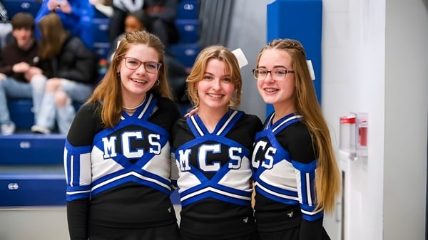 Tres estudiantes en uniformes de animación con el logo de MCS posan frente a un fondo azul y blanco en el campus de la Middletown Christian School.