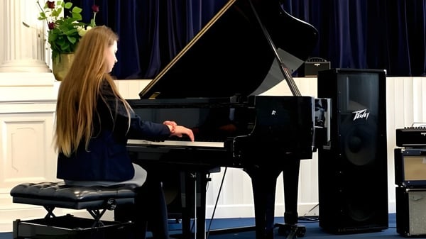 Una mujer con cabello largo y rubio toca el piano en el escenario del Midleton College frente a una cortina negra.