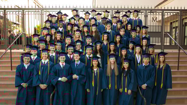 Un gran grupo de graduados está de pie en togas azules y doradas en los escalones de un edificio en el campus de la Mission College Preparatory Catholic High School.