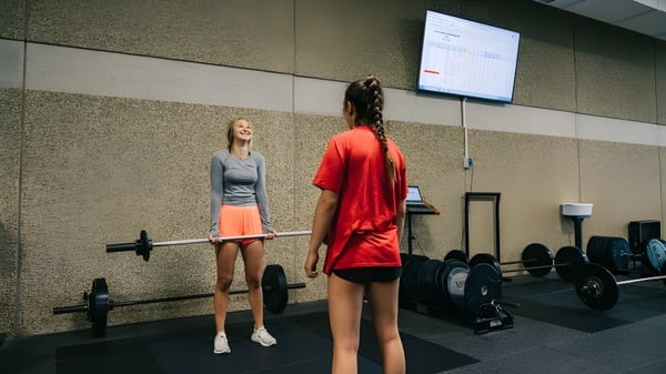 Dos mujeres en ropa deportiva están en el gimnasio de la Mission Secondary School frente a equipos de entrenamiento y una pantalla.