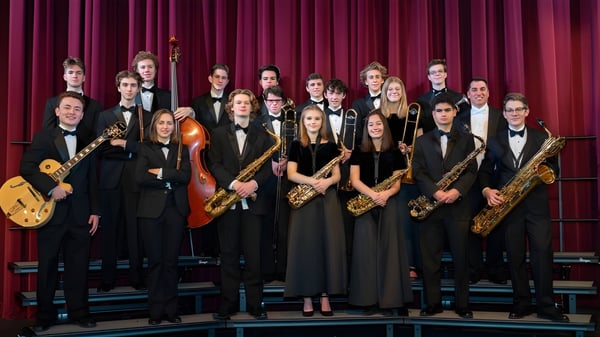 Un grupo de músicos del Moate Community College está vestido formalmente con instrumentos en un escenario frente a una cortina roja.