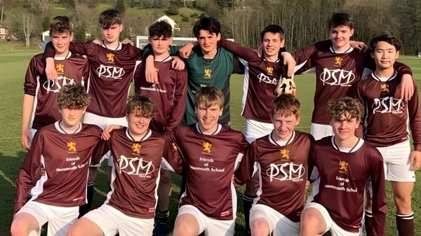 Un grupo de estudiantes de la Monmouth School for Boys posan en ropa deportiva sobre un campo de césped rodeado de árboles.