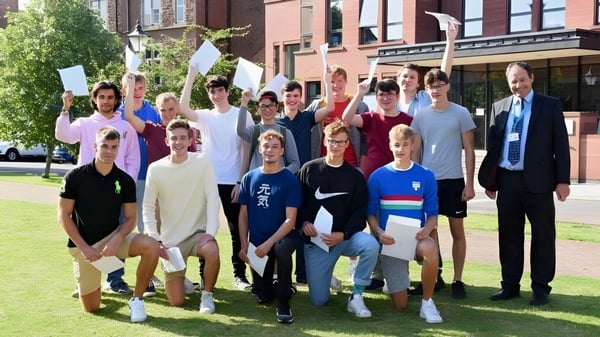 Estudiantes y adultos están juntos en un prado frente al edificio de ladrillo de la Monmouth School for Boys.