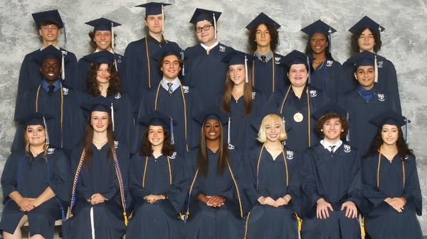Un grupo de graduados de la Monsignor Donovan Catholic High School de pie juntos en togas académicas frente a un fondo gris.
