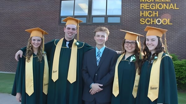 Un grupo de graduadas y graduados de la Morell Regional High School en togas verdes se encuentra frente al edificio escolar con un administrador escolar.