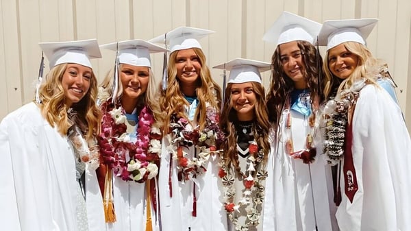 Un grupo de jóvenes graduadas de la Morgan High School se encuentra en ropa de graduación frente a una pared de madera.