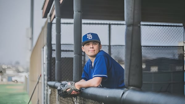 Un joven jugador de béisbol de la Morro Bay High School está en uniforme azul detrás de una cerca de alambre con un campo de béisbol al fondo.