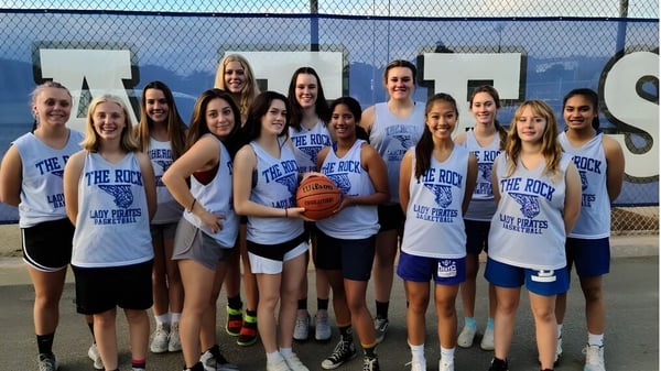 Un grupo de jóvenes jugadoras de baloncesto de la Morro Bay High School está juntas en un patio exterior con vista a la ciudad.