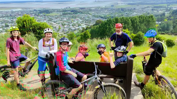 Un grupo de estudiantes de la Motueka High School en colorido equipo de ciclismo posan en un sendero montañoso con vista a la ciudad al fondo.