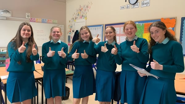 Seis estudiantes en uniformes idénticos están en un aula colorida en el campus de la Mount Anville Secondary School y muestran el pulgar hacia arriba.
