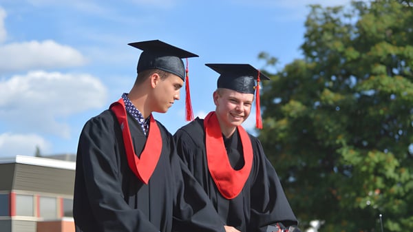 Dos estudiantes de la Mount Baker Secondary School están en sus togas de graduación al aire libre frente a árboles y un cielo azul.