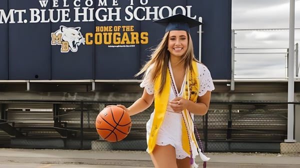 Una graduada de la Mount Blue High School está en un traje de gala y con un balón de baloncesto frente a un letrero escolar.