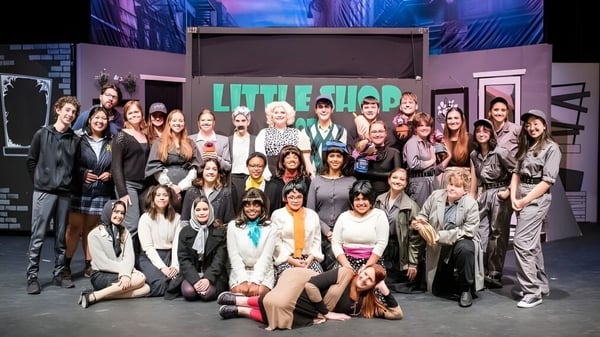 Un grupo de estudiantes de la Mount De Sales Academy posan juntos frente a un colorido fondo urbano con la inscripción Little Shop.