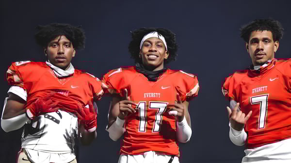 Tres estudiantes de la Mount Everett Regional High School llevan camisetas rojas de fútbol americano con los números 17 y 7 frente a un fondo oscuro.