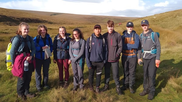 Un grupo de estudiantes de la Mount Kelly School está al aire libre en un paisaje montañoso bajo un cielo nublado.