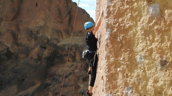 Un estudiante de la Mount Boucherie Secondary School escala con equipo en una empinada pared de roca en un entorno natural.