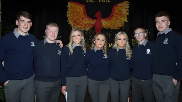 Un grupo de alumnos de la Mountmellick Community School está frente a un gran emblema parecido a un fénix sobre un fondo oscuro.