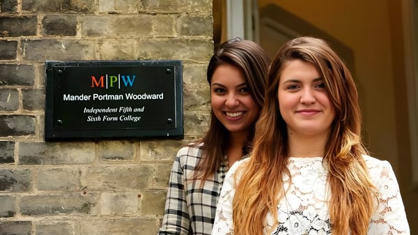 Dos estudiantes con cabello largo y oscuro están frente a una pared de ladrillo con el letrero de MPW Cambridge.