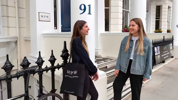 Dos estudiantes están de pie frente a un edificio con el número 91 en el campus de la MPW London y están conversando.