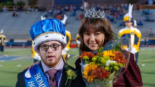 Un hombre con un sombrero azul y una mujer con una corona sostienen flores en un evento del Mt. Lebanon School District.