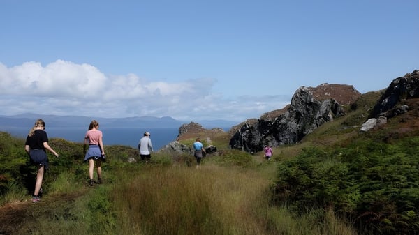 Estudiantes del Mullingar Community College caminan por un sendero a través de un paisaje verde con montañas y agua de fondo.