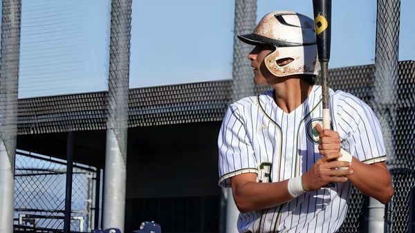 Un jugador de béisbol con uniforme a rayas blanco y azul sostiene un bate frente a una estructura similar a un estadio en el terreno del Murrieta Valley Unified School District.