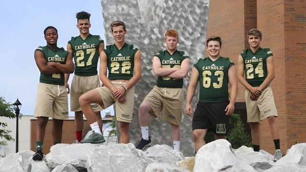Un grupo de deportistas está de pie en un saliente rocoso frente a un edificio en el campus de la Muskegon Catholic Central High School.